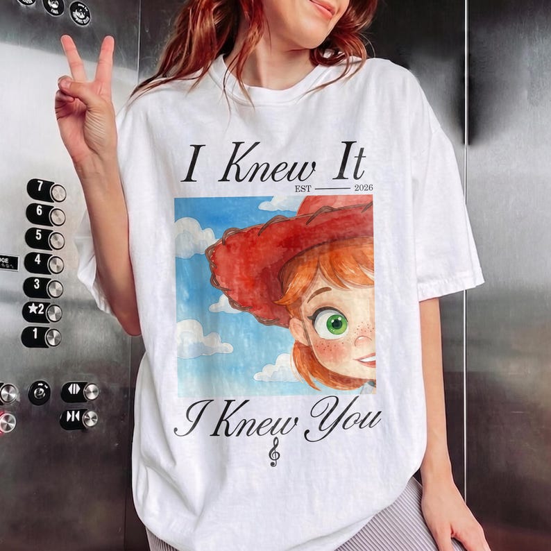 K&ouml;nnte beinhalten: Ein wei&szlig;es T-Shirt mit kurzen &Auml;rmeln und einem Grafikdruck einer Zeichentrickfigur mit roten Haaren und einem roten Hut vor einem blauen Himmel mit Wolken. Der Text lautet I Knew It EST 2026 oben und I Knew You mit einem Violinschl&uuml;ssel unten.