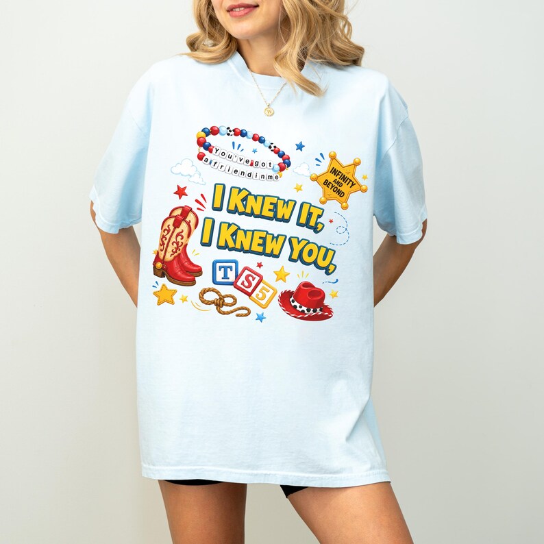 May include: A light blue t-shirt featuring a graphic print with the text "I KNEW IT, I KNEW YOU," a beaded bracelet, a sheriff badge with "INFINITY AND BEYOND," cowboy boots, "TS5" blocks, a lasso, and a red cowboy hat with white spots.