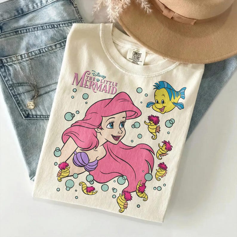 May include: Cream-colored t-shirt featuring a graphic of Ariel from Disney's The Little Mermaid. The design includes Ariel with pink hair, a purple seashell bra, and Flounder. The shirt also has seahorse and bubble illustrations.