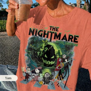 May include: Coral-colored t-shirt featuring a graphic from "The Nightmare Before Christmas." The design includes the film's title, Jack Skellington, Sally, and other characters in a spooky, Halloween-themed illustration. The shirt is short-sleeved.