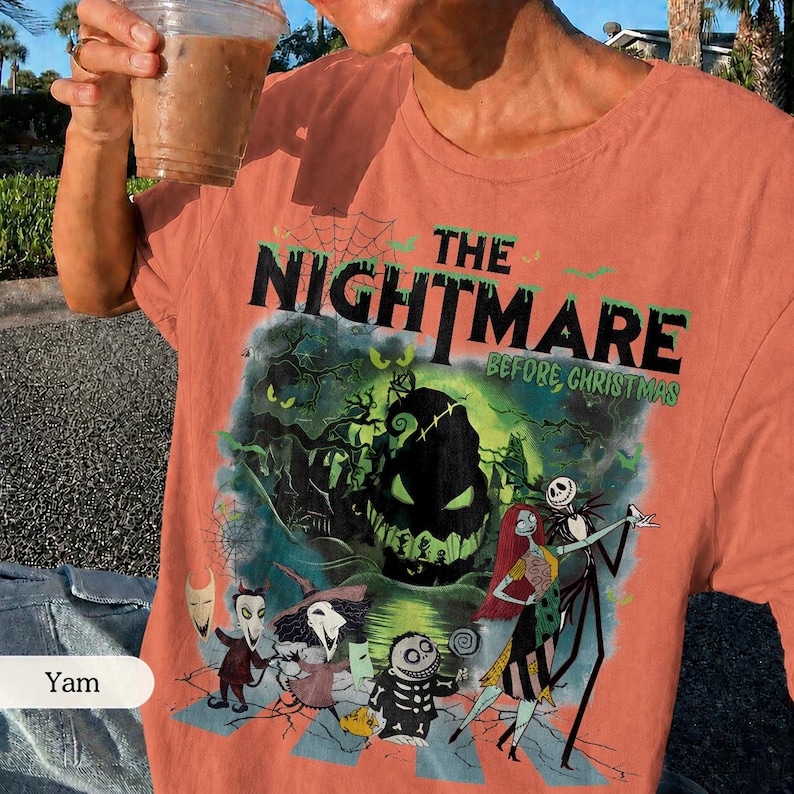 May include: Coral-colored t-shirt featuring a graphic from "The Nightmare Before Christmas." The design includes the film's title, Jack Skellington, Sally, and other characters in a spooky, Halloween-themed illustration. The shirt is short-sleeved.