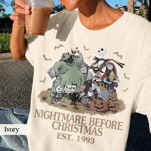May include: Ivory-colored t-shirt featuring a graphic from "The Nightmare Before Christmas." The design includes characters like Jack Skellington, Sally, and Oogie Boogie, with pumpkins and bats. The text "NIGHTMARE BEFORE CHRISTMAS EST. 1993" is printed below the graphic.