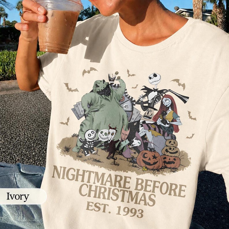 May include: Ivory-colored t-shirt featuring a graphic from "The Nightmare Before Christmas." The design includes characters like Jack Skellington, Sally, and Oogie Boogie, with pumpkins and bats. The text "NIGHTMARE BEFORE CHRISTMAS EST. 1993" is printed below the graphic.