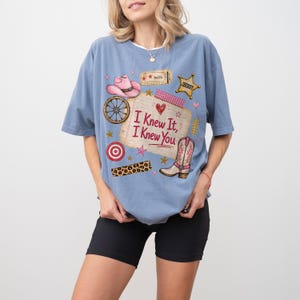 May include: A dusty blue short-sleeve t-shirt featuring a graphic print of a lined notepad with the text "I Knew It, I Knew You" surrounded by illustrations of a pink cowboy hat, a wagon wheel, a sheriff star, a target, and a cowboy boot.