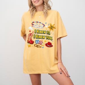 May include: A yellow short-sleeve t-shirt featuring a graphic print with the text I KNEW IT, I KNEW YOU, TS5, and INFINITY AND BEYOND. The design includes a beaded bracelet, a sheriff star, a cowboy boot, alphabet blocks, a lasso, and a cowboy hat.
