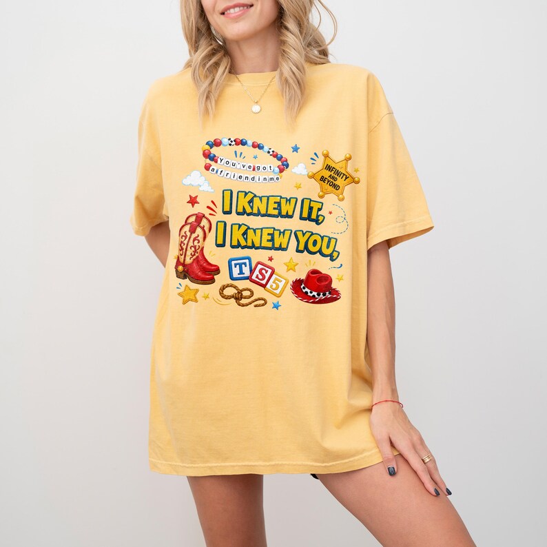 May include: A yellow short-sleeve t-shirt featuring a graphic print with the text I KNEW IT, I KNEW YOU, TS5, and INFINITY AND BEYOND. The design includes a beaded bracelet, a sheriff star, a cowboy boot, alphabet blocks, a lasso, and a cowboy hat.