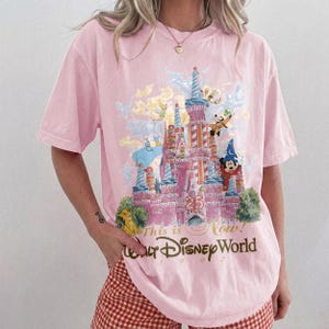 May include: A light pink short-sleeve t-shirt featuring a graphic of a castle with the number 25, surrounded by various cartoon characters. The text reads "This is the Magic" and "Walt Disney World 1997" in a stylized brown script font.