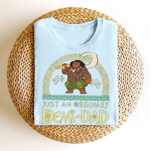 Puede incluir: Camiseta azul claro con una ilustraci&oacute;n de dibujos animados de un personaje con tatuajes, sosteniendo un arma y usando una falda de hierba. El texto "JUST AN ORDINARY DEMI-DAD" est&aacute; debajo en amarillo.