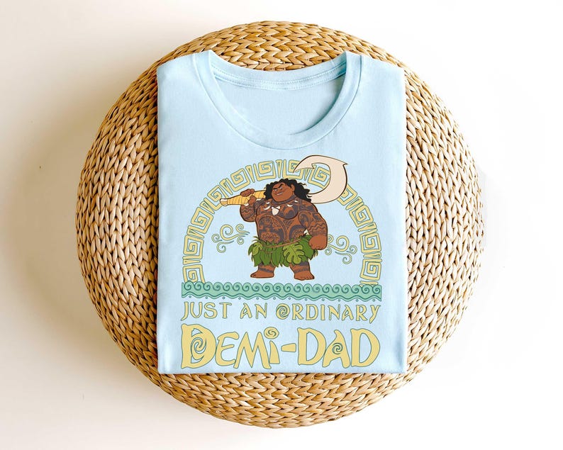 Puede incluir: Camiseta azul claro con una ilustraci&oacute;n de dibujos animados de un personaje con tatuajes, sosteniendo un arma y usando una falda de hierba. El texto "JUST AN ORDINARY DEMI-DAD" est&aacute; debajo en amarillo.