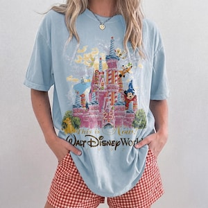May include: A light blue short-sleeve t-shirt featuring a graphic of a pink castle surrounded by various cartoon characters. The text reads This is Now! Walt Disney World 1997. The design includes a large central castle with a 25th anniversary emblem.
