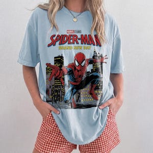 May include: A light blue short-sleeve t-shirt featuring a graphic print of Spider-Man in a city setting. The text reads Marvel Studios Spider-Man Brand New Day. The shirt has a crew neckline and a relaxed fit, showcasing a red and blue superhero.