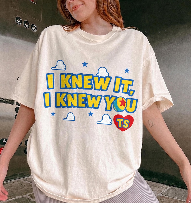 May include: A cream-colored short-sleeve t-shirt featuring the blue and yellow text I KNEW IT, I KNEW YOU with a colorful ball graphic, a red heart containing the letters TS, and small blue cloud and star icons printed on the front chest.