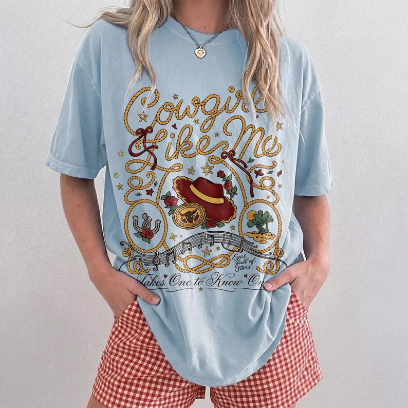 May include: A light blue t-shirt featuring a graphic print with the text Cowgirl Like Me, Eyes full of Stars, and Takes One to Know One. The design includes a red cowboy hat, a gold belt buckle, a horseshoe, a cactus, and rope elements.