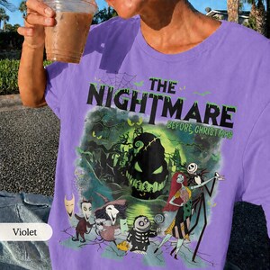 May include: Violet-colored t-shirt featuring a graphic from "The Nightmare Before Christmas." The design includes characters like Jack Skellington and Sally, with the movie title in green. The shirt is a crew neck.