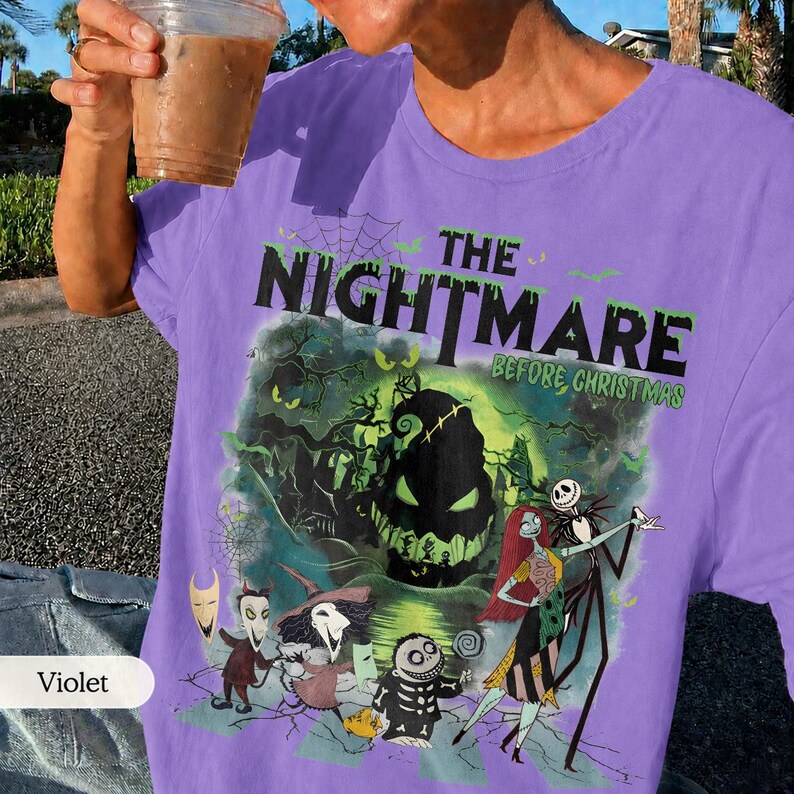 May include: Violet-colored t-shirt featuring a graphic from "The Nightmare Before Christmas." The design includes characters like Jack Skellington and Sally, with the movie title in green. The shirt is a crew neck.