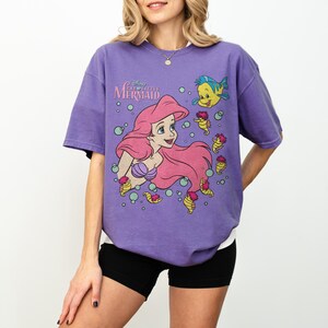 May include: Purple t-shirt featuring a graphic of Ariel from Disney's The Little Mermaid. The design includes Ariel with pink hair, a blue fish, and seahorses. The shirt has a crew neck and short sleeves.