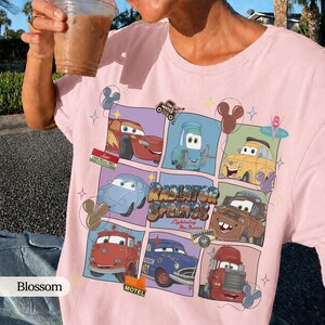 May include: A light pink t-shirt featuring a grid graphic of various characters from the movie Cars, including Lightning McQueen, Mater, and Sally. The design includes the text Radiator Springs and Lightning McQueen, surrounded by small star and Mickey icon shapes.