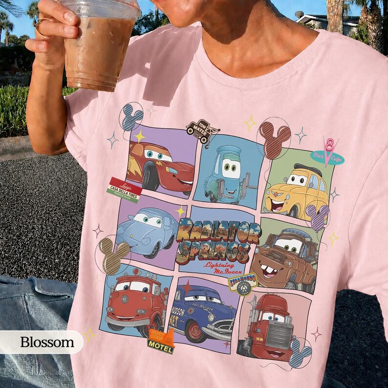 May include: A light pink t-shirt featuring a grid graphic of various characters from the movie Cars, including Lightning McQueen, Mater, and Sally. The design includes the text Radiator Springs and Lightning McQueen, surrounded by small star and Mickey icon shapes.