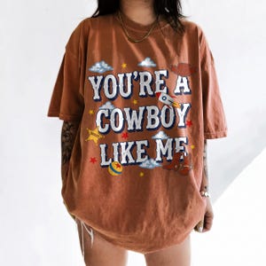 Puede incluir: Una camiseta de manga corta color &oacute;xido con el texto "YOU'RE A COWBOY LIKE ME" en blanco con bordes azules. El gr&aacute;fico incluye ilustraciones de un cohete, un sombrero de vaquero, botas, una estrella de sheriff, una pelota, nubes y estrellas.