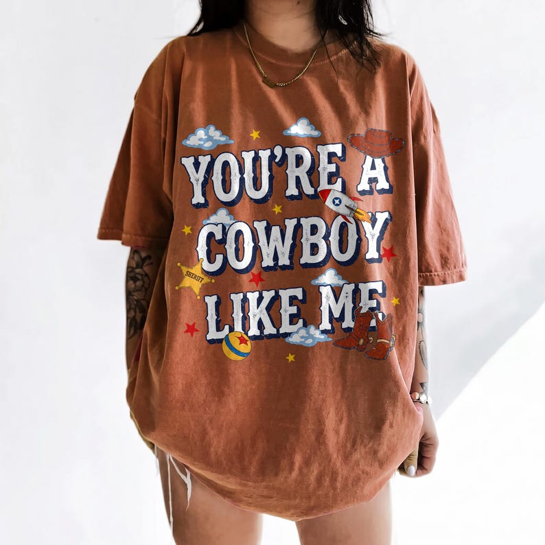 Puede incluir: Una camiseta de manga corta color &oacute;xido con el texto "YOU'RE A COWBOY LIKE ME" en blanco con bordes azules. El gr&aacute;fico incluye ilustraciones de un cohete, un sombrero de vaquero, botas, una estrella de sheriff, una pelota, nubes y estrellas.