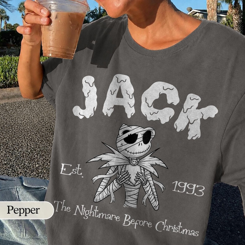 May include: Dark gray t-shirt featuring the word "JACK" in a dripping font above a mummy-like character. Below the character is the text "The Nightmare Before Christmas" and the year "1993".