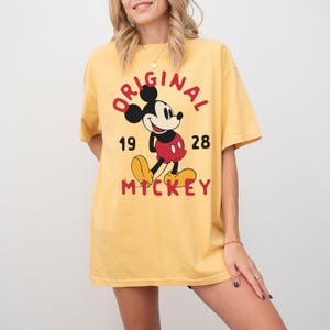 May include: Yellow t-shirt featuring a classic Mickey Mouse graphic. The design includes the word "ORIGINAL" in red, the year "1928", and the name "MICKEY" in red. The shirt is a casual, oversized fit.