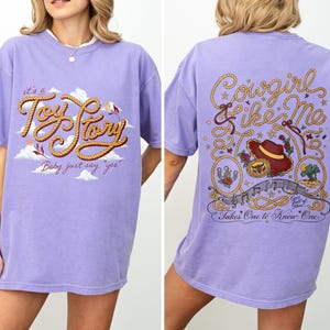 May include: A light purple t-shirt featuring a front graphic with the text "it's a Toy Story" and "Baby just say yes" with clouds. The back graphic displays the text "Cowgirl like Me" and "Takes One to Know One" with a hat, rope, and stars.