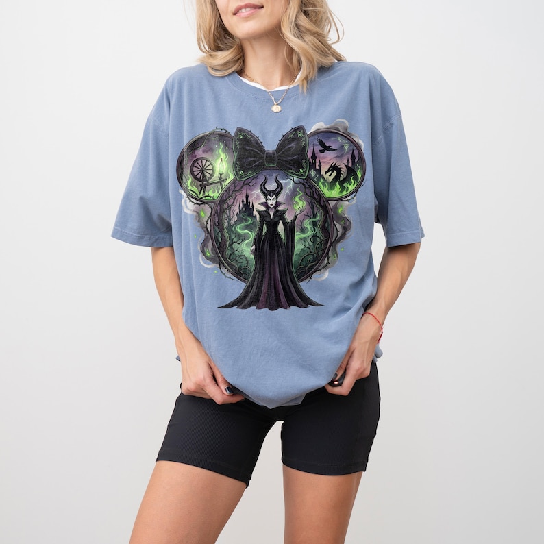 May include: Blue t-shirt featuring a graphic of a Disney villain in a black dress, surrounded by green and black imagery. The design is set within a Mickey Mouse ear shape, with a black bow at the top.