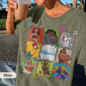 Op de afbeelding: Mosgroen t-shirt met een kleurrijke afbeelding van personages uit de Disney-film Moana. Het ontwerp bevat Moana, Maui, Pua, Hei Hei en andere personages. Het shirt heeft een ronde hals en korte mouwen.