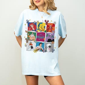 May include: Light blue t-shirt featuring a colorful grid of characters from "The Little Mermaid." The grid includes Ariel, Flounder, Sebastian, King Triton, Ursula, and others. The shirt has a relaxed fit and short sleeves.