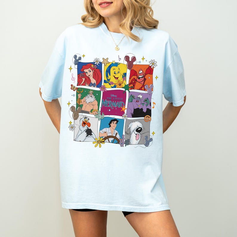 May include: Light blue t-shirt featuring a colorful grid of characters from "The Little Mermaid." The grid includes Ariel, Flounder, Sebastian, King Triton, Ursula, and others. The shirt has a relaxed fit and short sleeves.