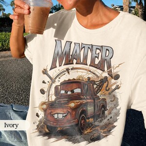 May include: Ivory-colored t-shirt featuring a graphic of Mater from the movie Cars. The design includes the name "MATER" in large letters above the character, along with wrenches and a speedometer. The cartoon tow truck is brown with a smiling face.