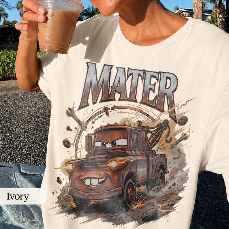 May include: Ivory-colored t-shirt featuring a graphic of Mater from the movie Cars. The design includes the name "MATER" in large letters above the character, along with wrenches and a speedometer. The cartoon tow truck is brown with a smiling face.