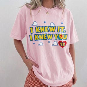 May include: A light pink short-sleeve t-shirt featuring the yellow text "I KNEW IT, I KNEW YOU" with a red heart containing the letters "TS" and a colorful ball graphic. The design includes small blue stars and white cloud outlines printed on the front.
