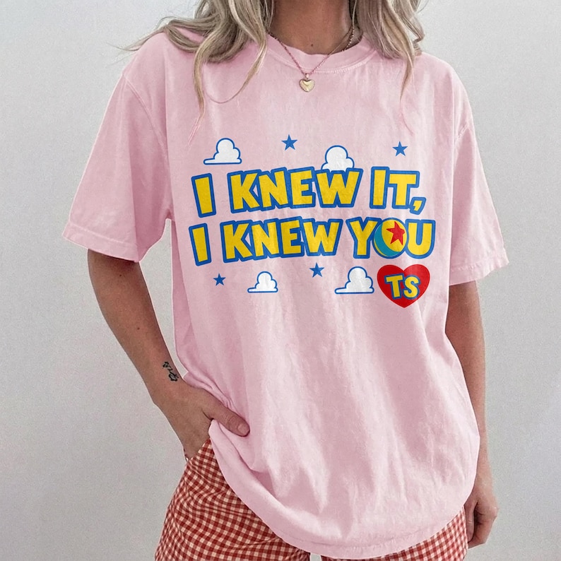 May include: A light pink short-sleeve t-shirt featuring the yellow text "I KNEW IT, I KNEW YOU" with a red heart containing the letters "TS" and a colorful ball graphic. The design includes small blue stars and white cloud outlines printed on the front.