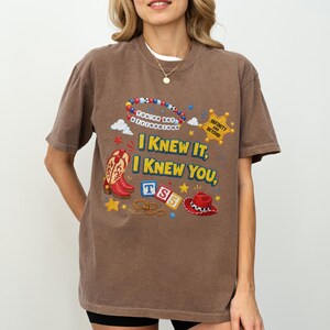 May include: A brown short-sleeve t-shirt featuring a graphic print with red cowboy boots, a red cowboy hat, a sheriff badge labeled "INFINITY AND BEYOND," and letter blocks spelling "TS5." The text reads "I KNEW IT, I KNEW YOU," and "You've got a friend in me."