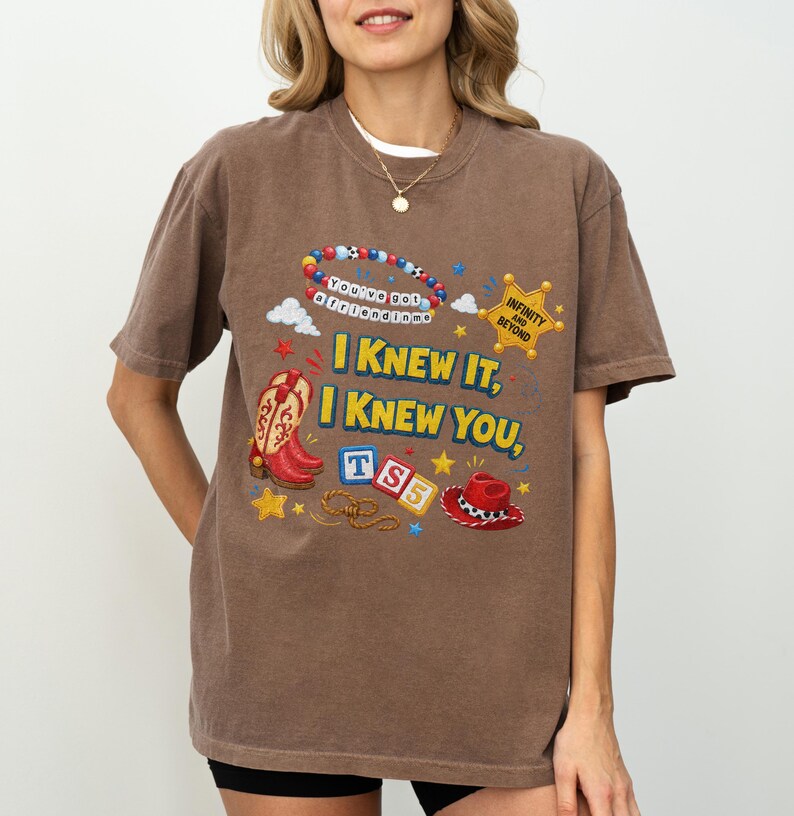May include: A brown short-sleeve t-shirt featuring a graphic print with red cowboy boots, a red cowboy hat, a sheriff badge labeled "INFINITY AND BEYOND," and letter blocks spelling "TS5." The text reads "I KNEW IT, I KNEW YOU," and "You've got a friend in me."