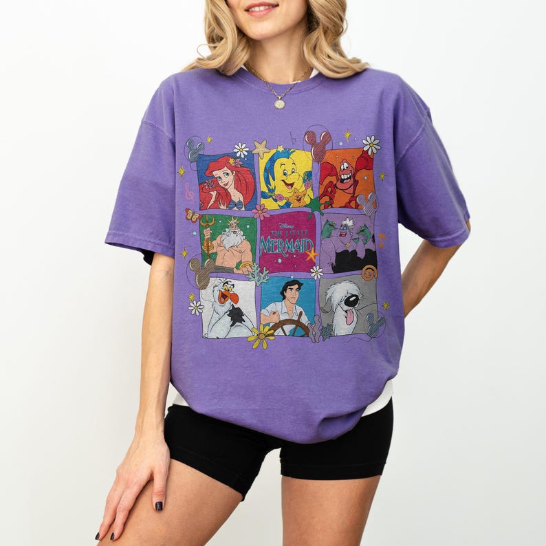 May include: Purple t-shirt featuring a colorful graphic of characters from Disney's The Little Mermaid. The design includes Ariel, Sebastian, Flounder, and other characters in a grid pattern. The shirt has short sleeves and a crew neck.