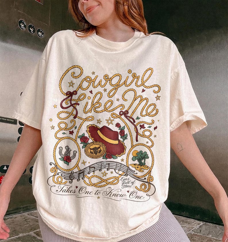 May include: An off-white short-sleeve t-shirt featuring a graphic print of a rope border, a cowboy hat, a horseshoe, a cactus, and musical notes. The text reads Cowgirl like Me and Takes One to Know One with the phrase Eyes full of Stars.