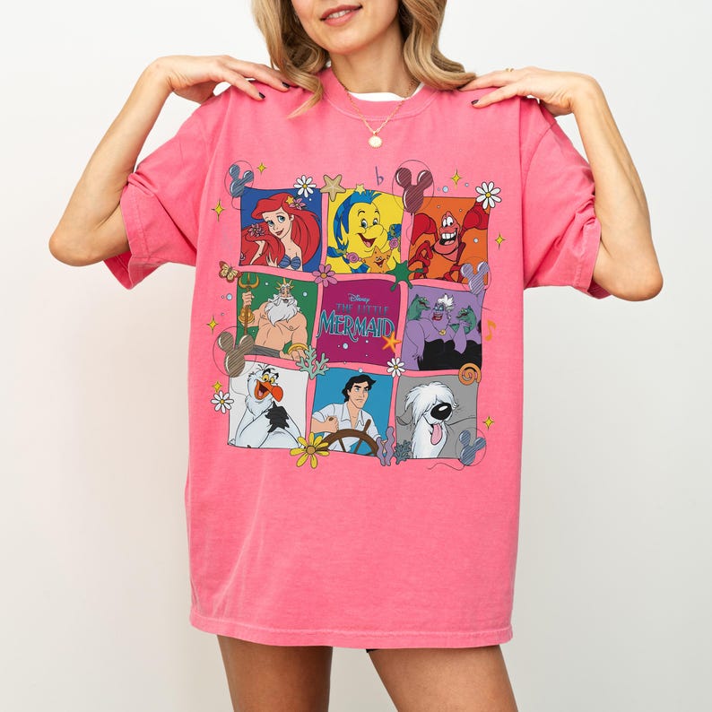 May include: Pink t-shirt featuring a grid of colorful illustrations of characters from Disney's "The Little Mermaid." The shirt includes Ariel, Flounder, Sebastian, King Triton, Ursula, Olaf, Prince Eric, and Max. The words "The Little Mermaid" are also displayed.