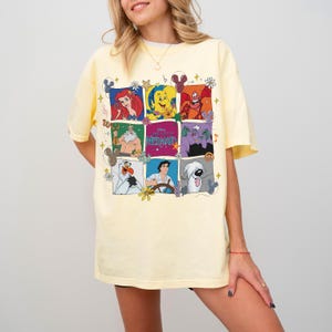 May include: A light yellow t-shirt featuring a colorful grid of characters from "The Little Mermaid." The grid includes Ariel, Flounder, Sebastian, King Triton, Ursula, Olaf, Prince Eric, and Max. The shirt has short sleeves and a crew neck.