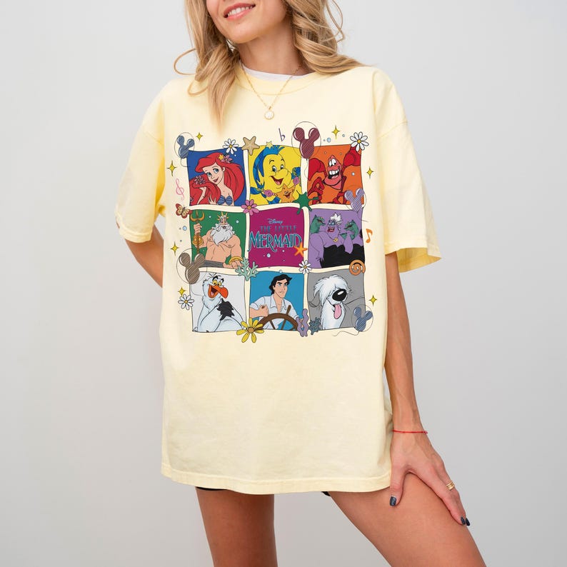 May include: A light yellow t-shirt featuring a colorful grid of characters from "The Little Mermaid." The grid includes Ariel, Flounder, Sebastian, King Triton, Ursula, Olaf, Prince Eric, and Max. The shirt has short sleeves and a crew neck.