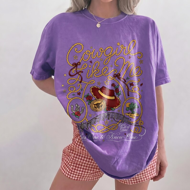 May include: A purple short-sleeve t-shirt featuring a graphic print of a yellow rope forming the text Cowgirl Like Me, a red cowboy hat, a horseshoe, a cactus, musical notes, and the text Eyes full of Stars... Takes One to Know One.