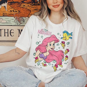May include: White t-shirt featuring a graphic of Ariel from Disney's The Little Mermaid. The design includes Ariel with pink hair, a purple seashell bra, and Flounder. The shirt has the text "The Little Mermaid".