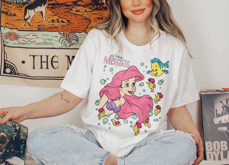 May include: White t-shirt featuring a graphic of Ariel from Disney's The Little Mermaid. The design includes Ariel with pink hair, a purple seashell bra, and Flounder. The shirt has the text "The Little Mermaid".