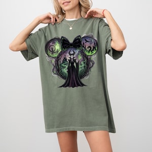 May include: Sage green t-shirt featuring a graphic of a character in a black dress with a bow, surrounded by a forest scene and a dragon. The design is set within a Mickey Mouse ear shape.