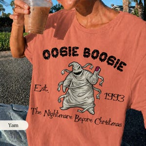 May include: Coral-colored t-shirt featuring a graphic of Oogie Boogie from "The Nightmare Before Christmas." The design includes the text "Oogie Boogie," "Est. 1993," and "The Nightmare Before Christmas."