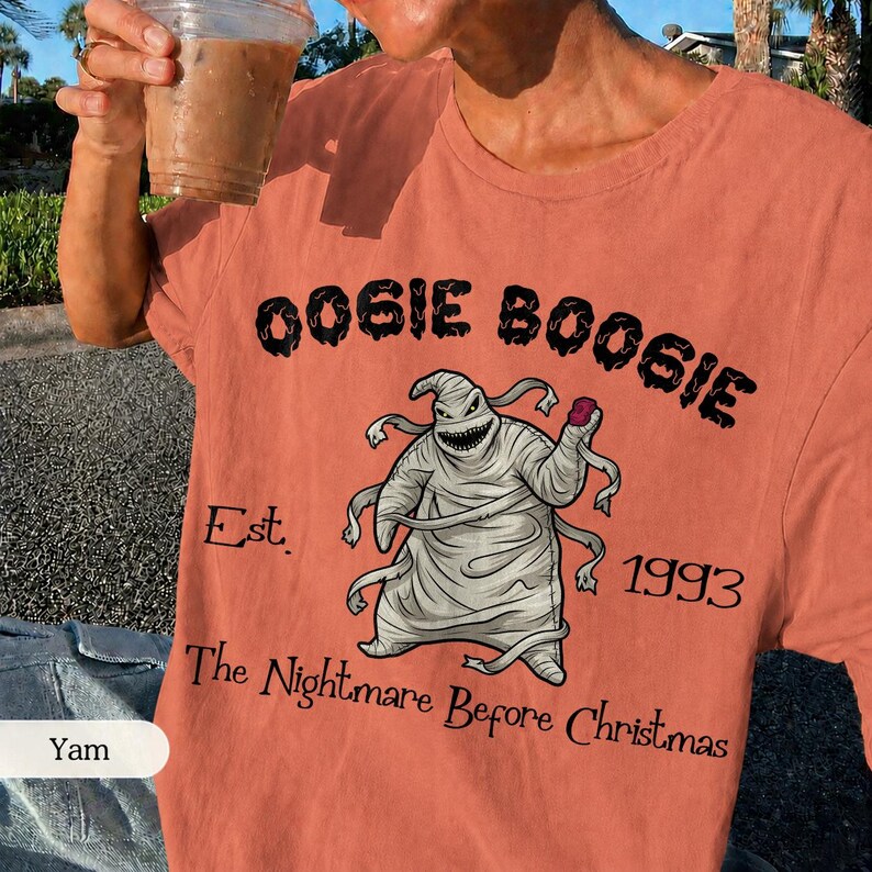 May include: Coral-colored t-shirt featuring a graphic of Oogie Boogie from "The Nightmare Before Christmas." The design includes the text "Oogie Boogie," "Est. 1993," and "The Nightmare Before Christmas."