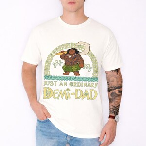Puede incluir: Camiseta de color crema con una ilustraci&oacute;n de dibujos animados de una figura polinesia sosteniendo un arma. El texto "JUST AN ORDINARY DEMI-DAD" est&aacute; debajo de la ilustraci&oacute;n en amarillo. La camiseta es de cuello redondo.