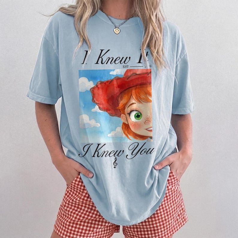 K&ouml;nnte beinhalten: Ein hellblaues Kurzarm-T-Shirt mit einer Grafik einer rothaarigen Figur mit rotem Hut vor einem blauen Himmel mit Wolken. Der Text lautet I Knew It EST 2016 und I Knew You, erg&auml;nzt durch ein Violinschl&uuml;ssel-Symbol unter dem Text.